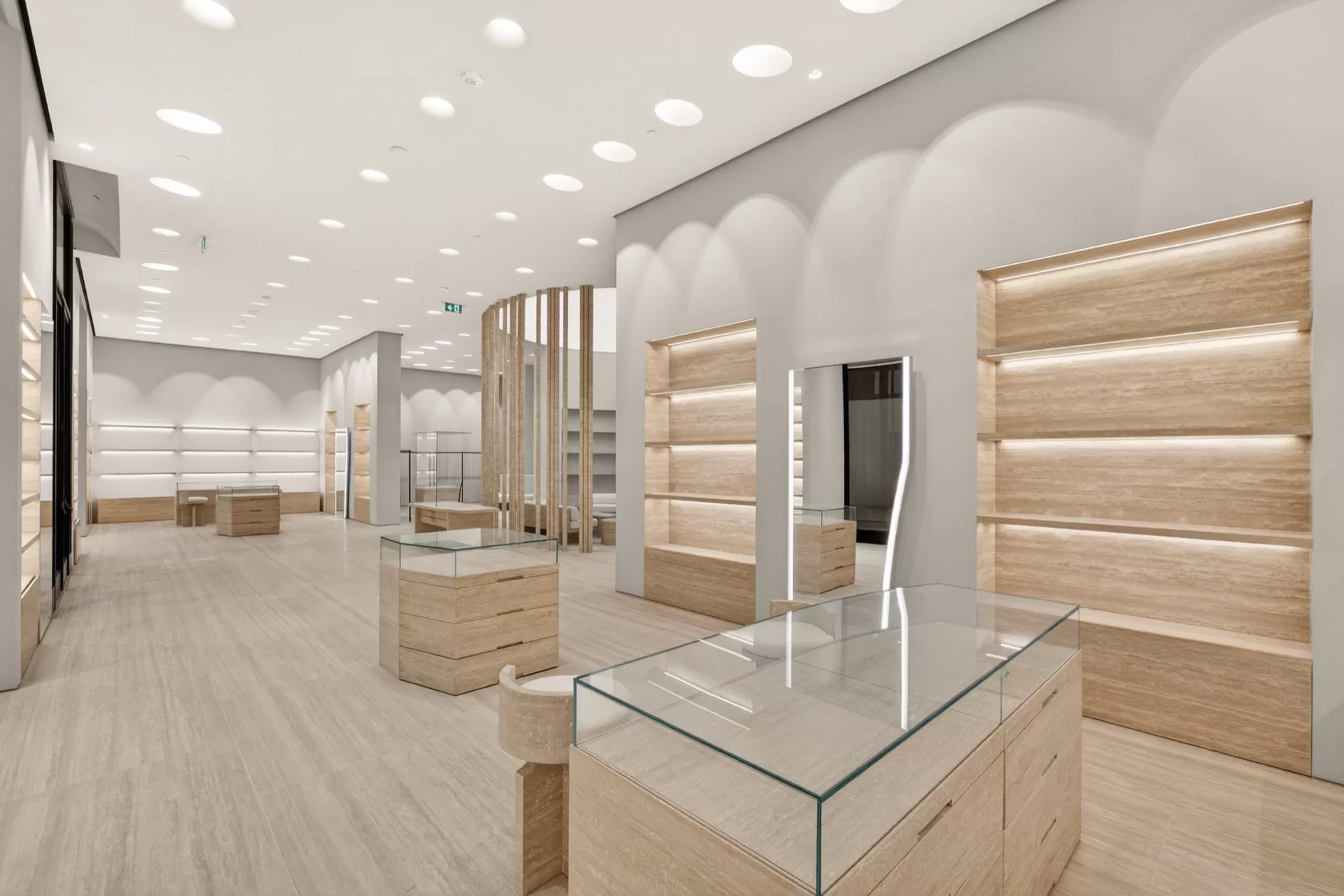 Maison Margiela Oakridge luxury retail sales floor — travertine display plinths with glass vitrines and illuminated wall shelving beneath arched white plaster niches