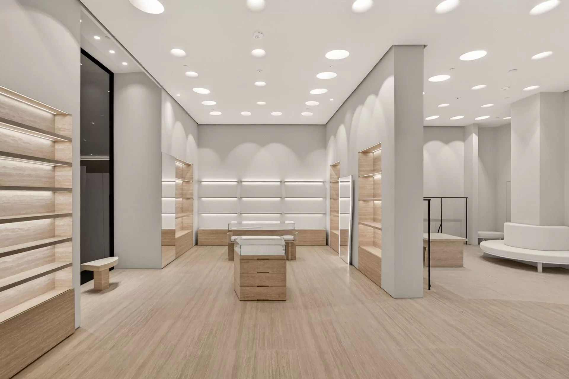 Luxury retail display hall — backlit travertine wall shelving, central glass display cases and scalloped cove-lit white plaster ceilings